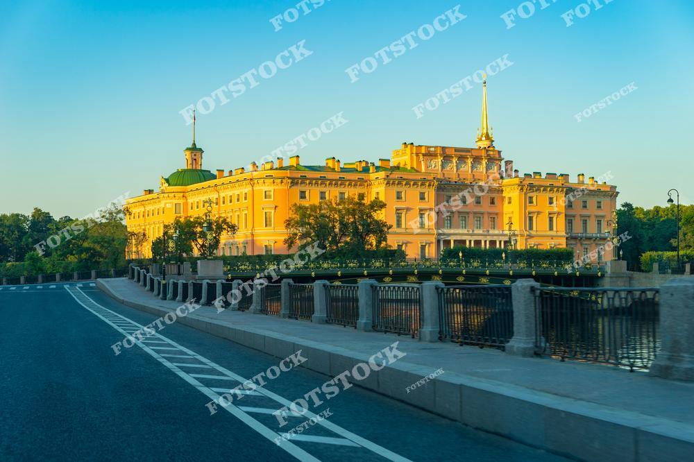 На фотографии изображен Михайловский замок — уникальное архитектурное сооружение в Санкт-Петербурге, построенное по приказу императора Павла I. Его яркий фасад и изящный шпиль выделяются на фоне ясного утреннего неба. Замок окружен каналом и мостами, придающими ему особую изолированность и величественность. Утренний свет делает здание еще более выразительным, подчеркивая его историческую значимость.